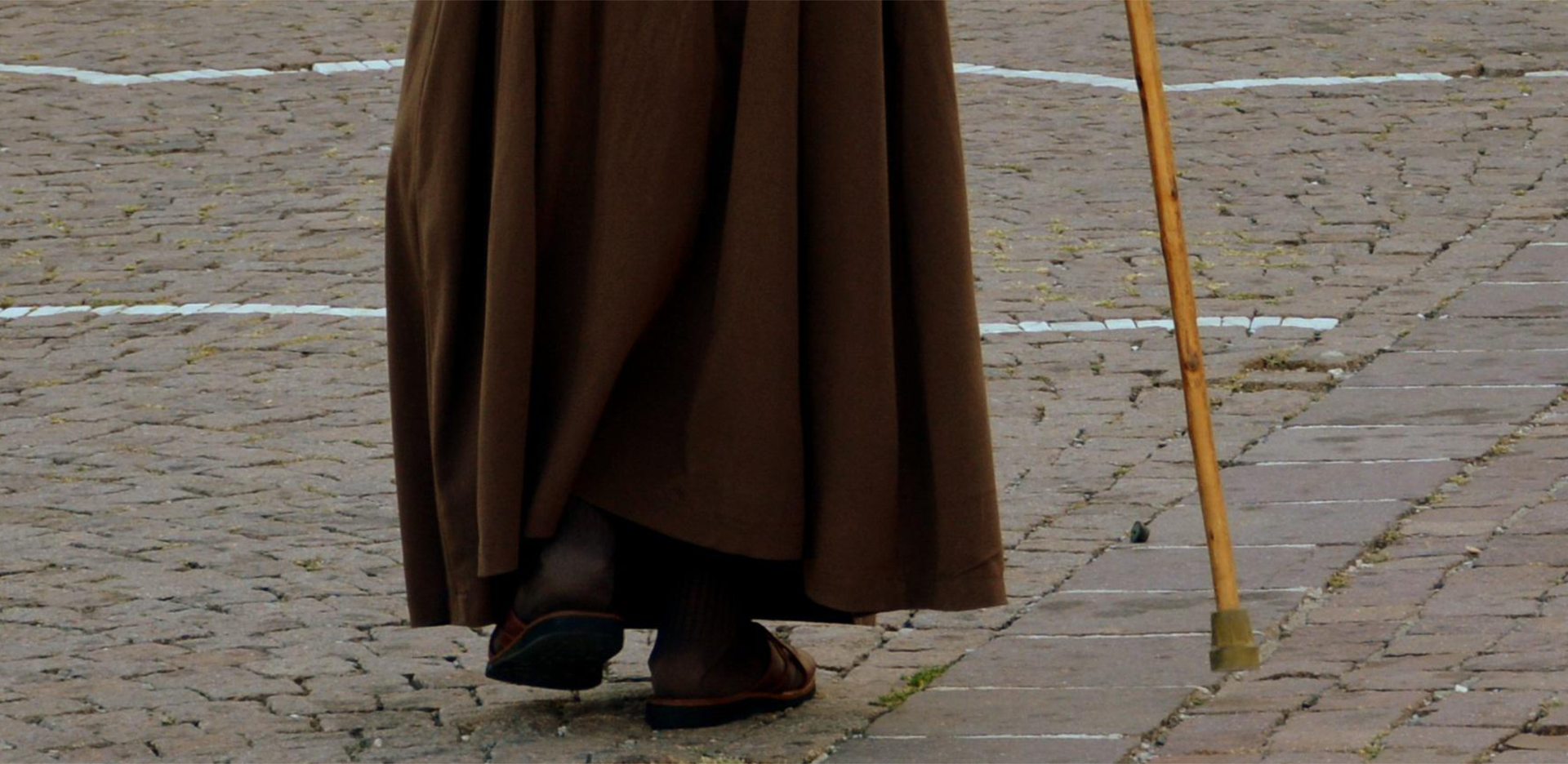 Zdjęcie przedstawia z tyłu nogi osoby w habicie idącej o lasce.