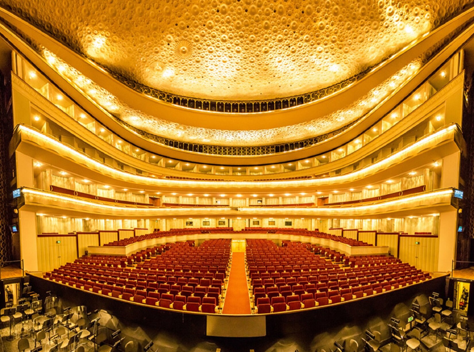 Zdjęcie przedstawia widownię w sali koncertowej. Widownia składa się z czerwonych foteli, ułożonych w równe rzędy. Pośrodku fotele przedzielone są korytarzem. Od lewej do prawej strony, na kilku poziomach znajdują się loże z innymi miejscami siedzącymi. Sufit i ściany sali mają jasny kolor.