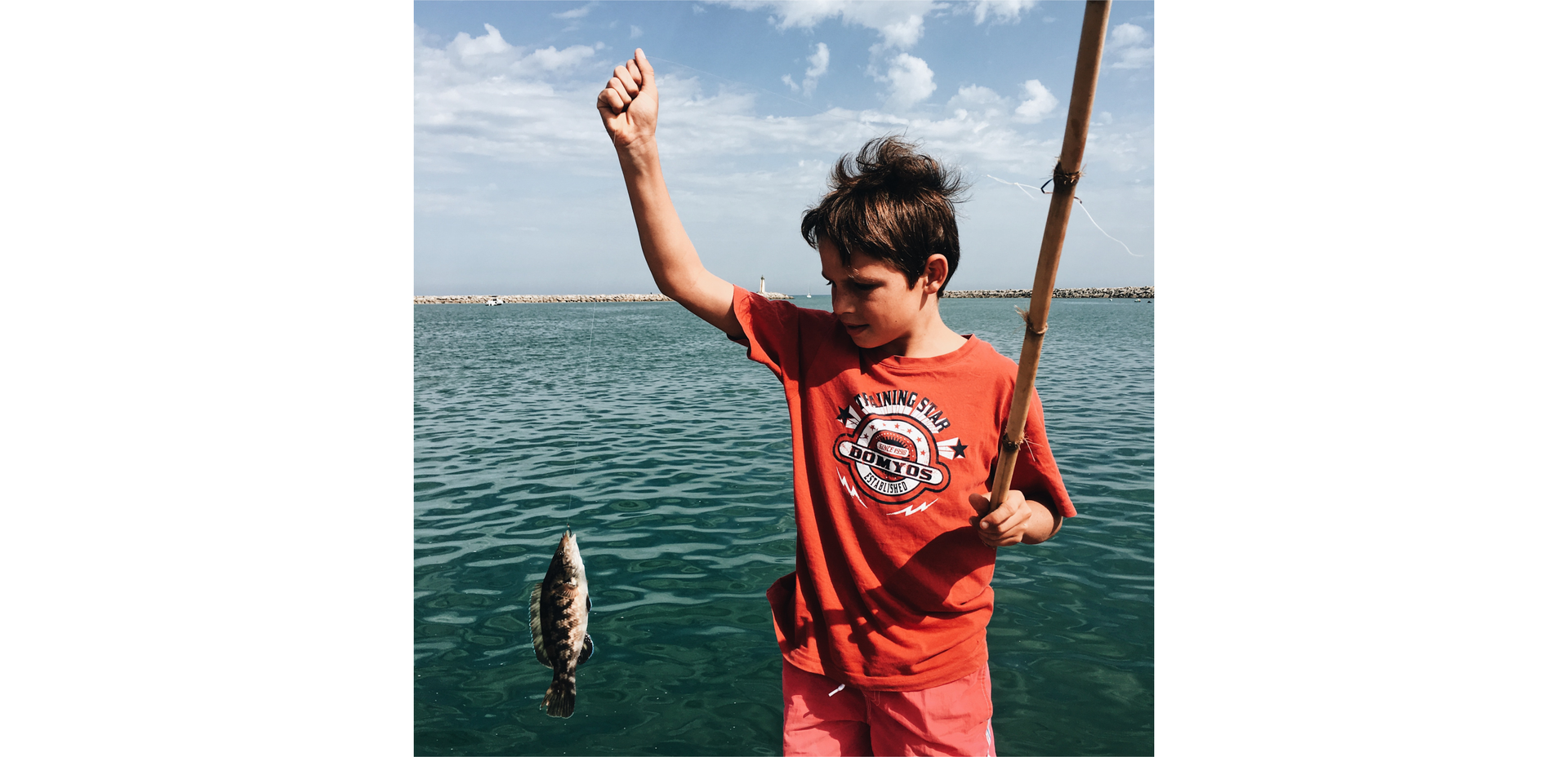 Zdjęcie przedstawia młodego chłopca, który trzyma w ręku żyłkę, na której wisi złowiona mała ryba.