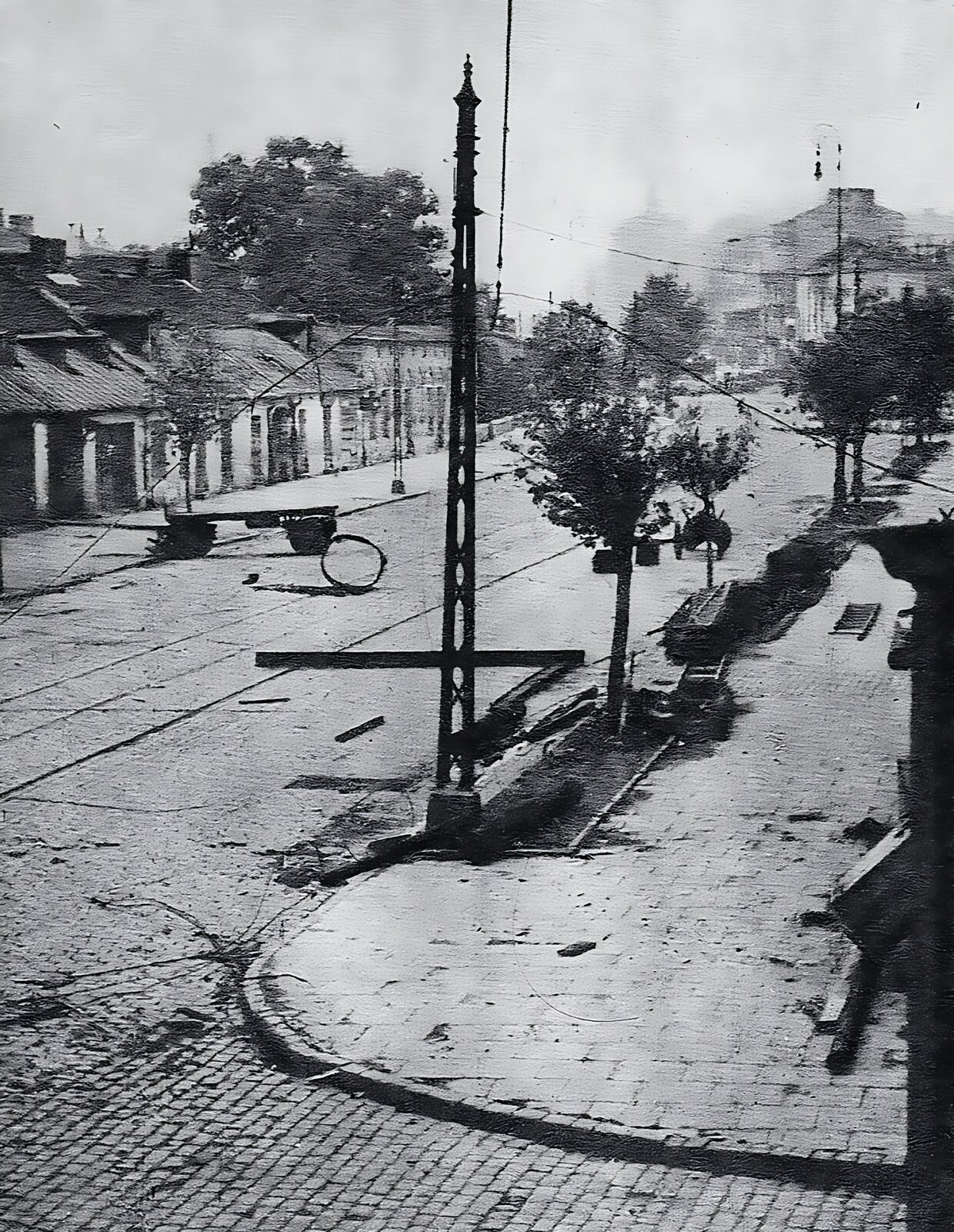 Stare zdjęcie przedstawia ulicę Wolską.  Ulica jest wybrukowana kostką kamienną.  Leżą na niej porozrzucane fragmenty wozu, szyny kolejowe. Na chodniku po prawej stronie kilka drabin. Widać niską zabudowę miejską. W głębi widoczne wyższe budynki. Wzdłuż ulicy rosną niewysokie drzewa.