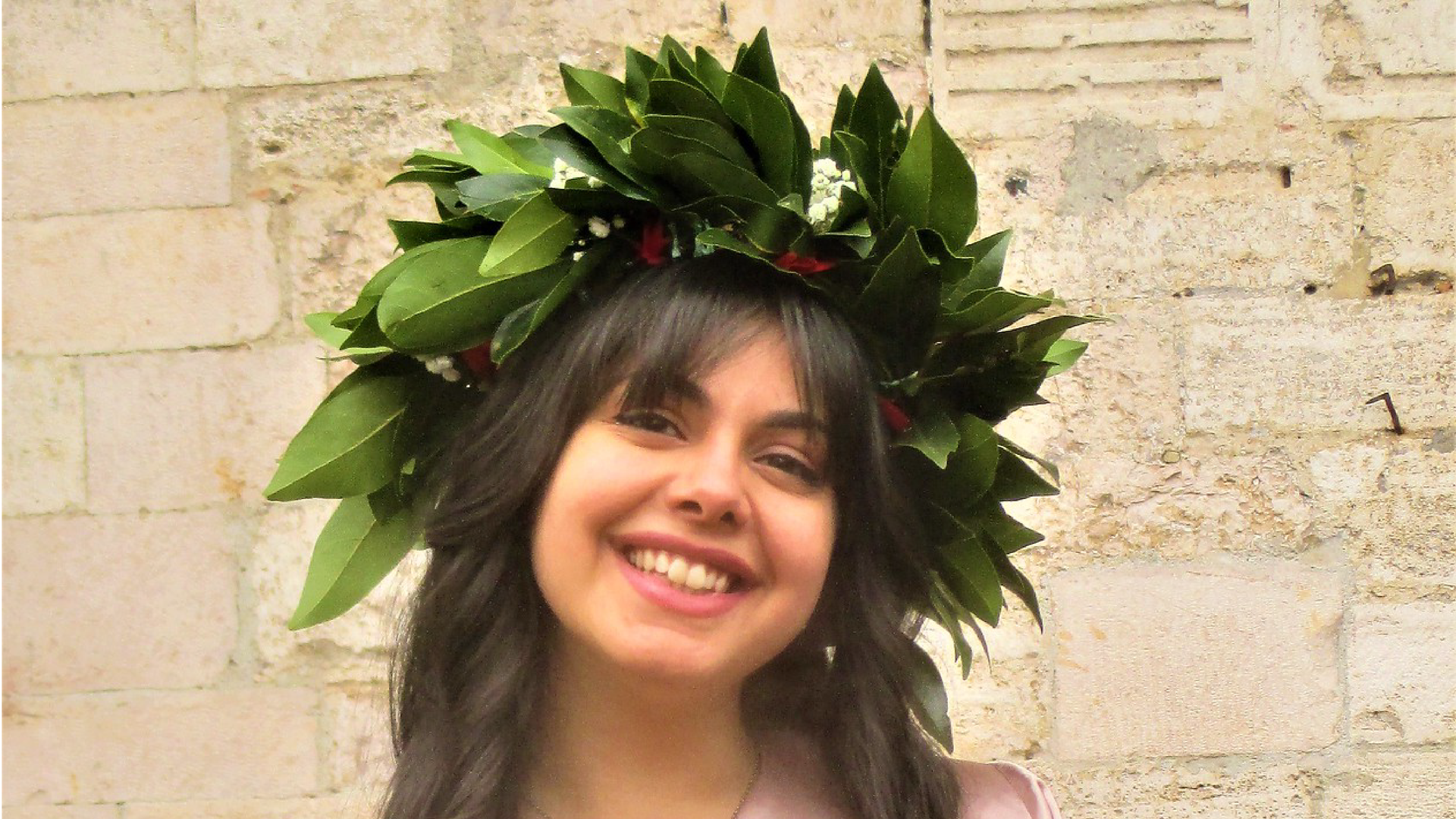 Zdjęcie przedstawia zbliżenie na twarz uśmiechniętej, młodej dziewczyny z długimi włosami. Dziewczyna ma na głowie wieniec laurowy.