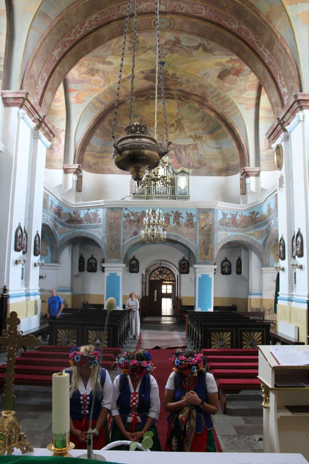 Zdjęcie przedstawia wnętrze kościoła z perspektywy ołtarza. Na pierwszym planie znajdują się trzy klękające kobiety, które obrane są w czerwone spódnice, białe koszule i niebieskie kamizelki. Na głowach mają kolorowe wianki. W tyłu, na kobietami, widoczne są czerwone ławki bez oparć, a za nimi ustawione zostały drewniane ławy z oparciami. Przy ławach stoją dwie osoby - kobieta w białym swetrze i spódnicy oraz łysy mężczyzna w niebieskiej koszulce. Za nimi widoczny jest chór, którego ściany zostały ozdobione licznymi malowidłami. Sufit kościoła również jest cały w malowidłach przedstawiających biblijne sceny. Na chórze stoją złoto‑niebieskie organy. Na pierwszym planie widoczny jest złoty pojemnik zawieszony na złotych łańcuchach.