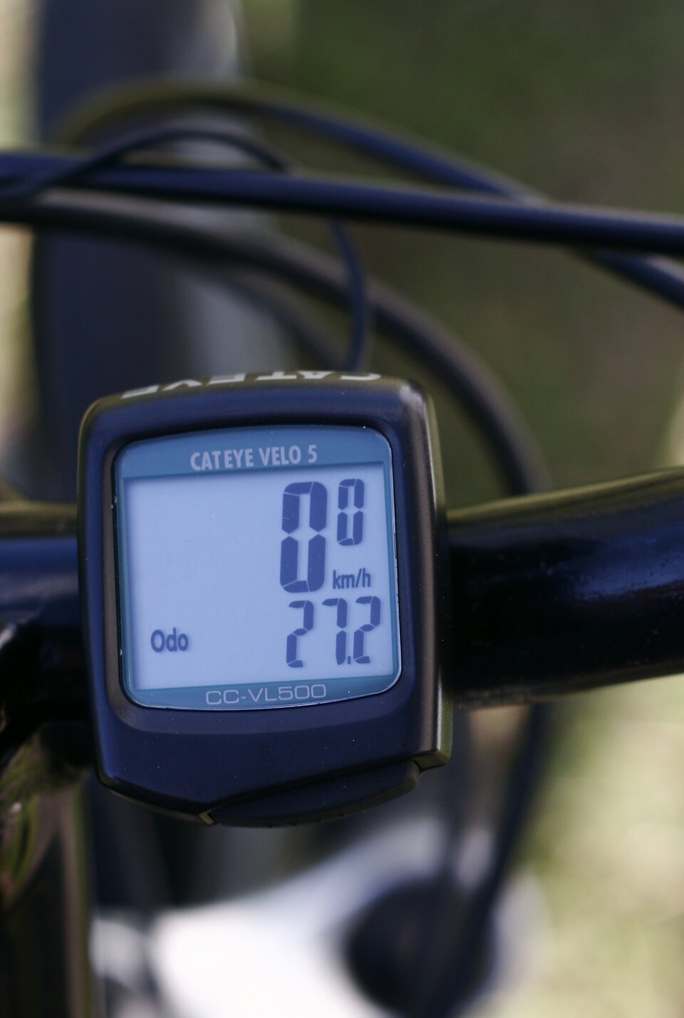 Zbliżenie na czarny, kwadratowy licznik rowerowy zamontowany na kierownicy. Na szarym wyświetlaczu widoczna jest aktualna prędkość zero kilometrów na godzinę w górnej części oraz przejechany dystans (27,2 kilometra) w dolnej części. Rower znajduje się w spoczynku.