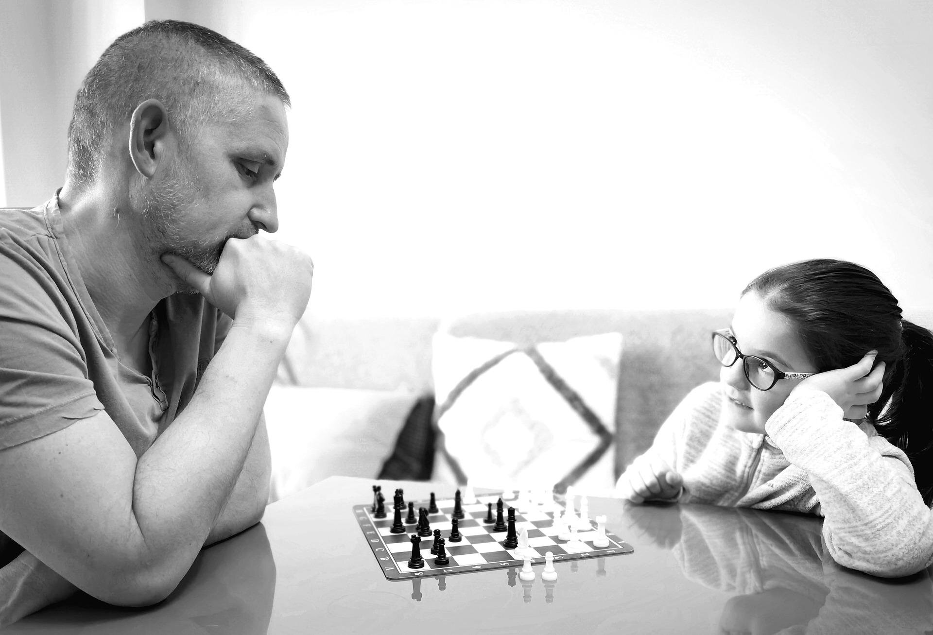 Zdjęcie przedstawia dziewczynkę i dorosłego mężczyznę grających w szachy. Mężczyzna ma skupiony wyraz twarzy.