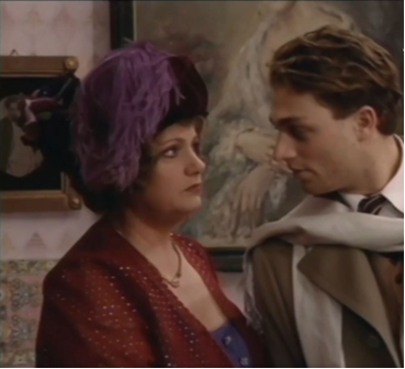 Zdjęcie przedstawia stojących obok siebie kobietę i mężczyznę, którzy patrzą sobie w oczy. Kobieta ma czerwoną bluzkę i ciemny kapelusz z fioletowymi piórami. Mężczyzna ma ciemne włosy i jasnobrązowy garnitur z białym szalem.