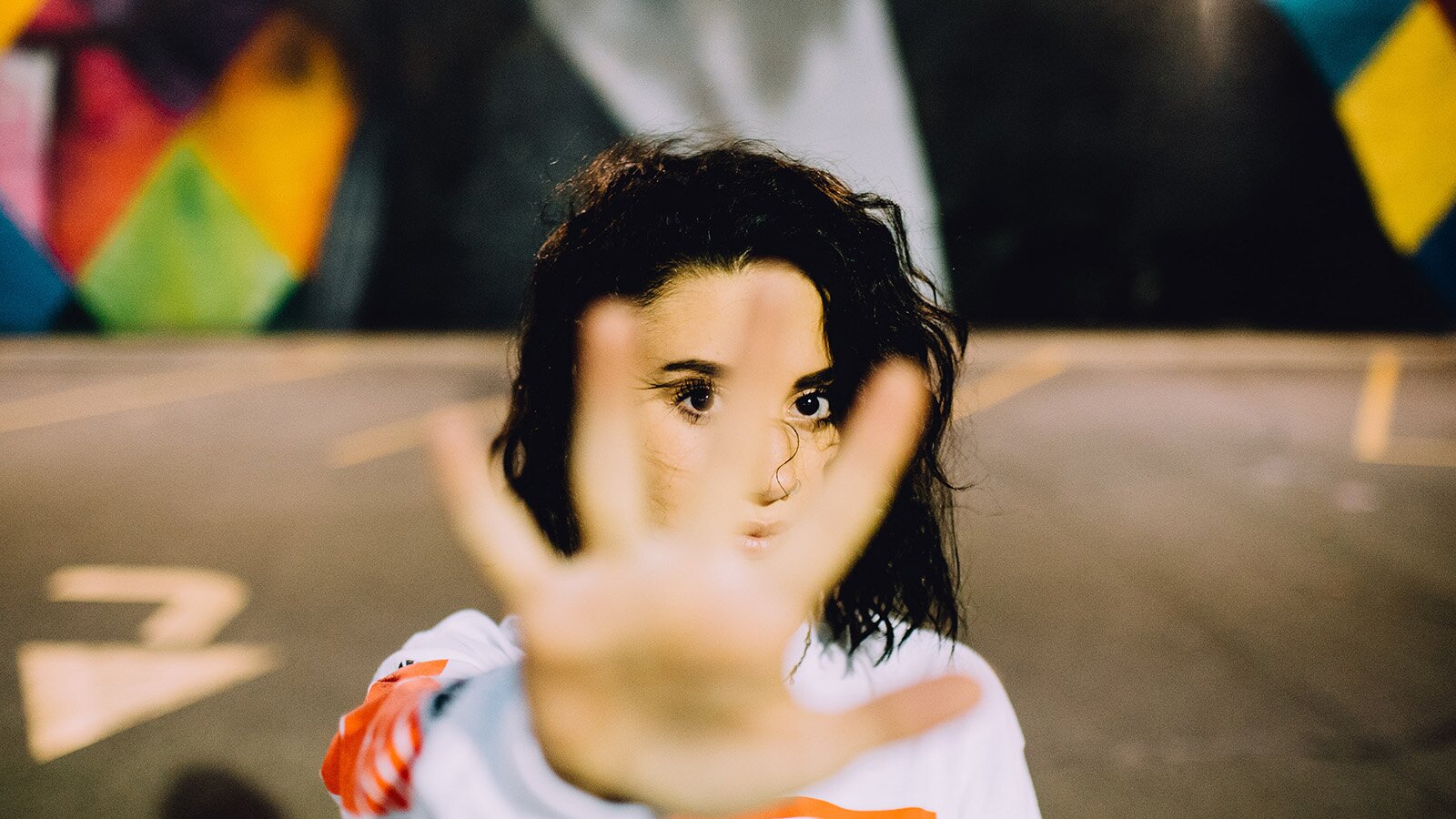 Zdjęcie przedstawia nastolatkę w ujęciu od klatki piersiowej w górę. Nastolatka ma wyciągniętą przed siebie prawą rękę, a jej dłoń pokazuje pięć palców. Nastolatka ma ciemne, rozpuszczone włosy i jest ubrana w jasną bluzę. W tle widoczna jest rozmyta podłoga oraz kolorowe elementy.