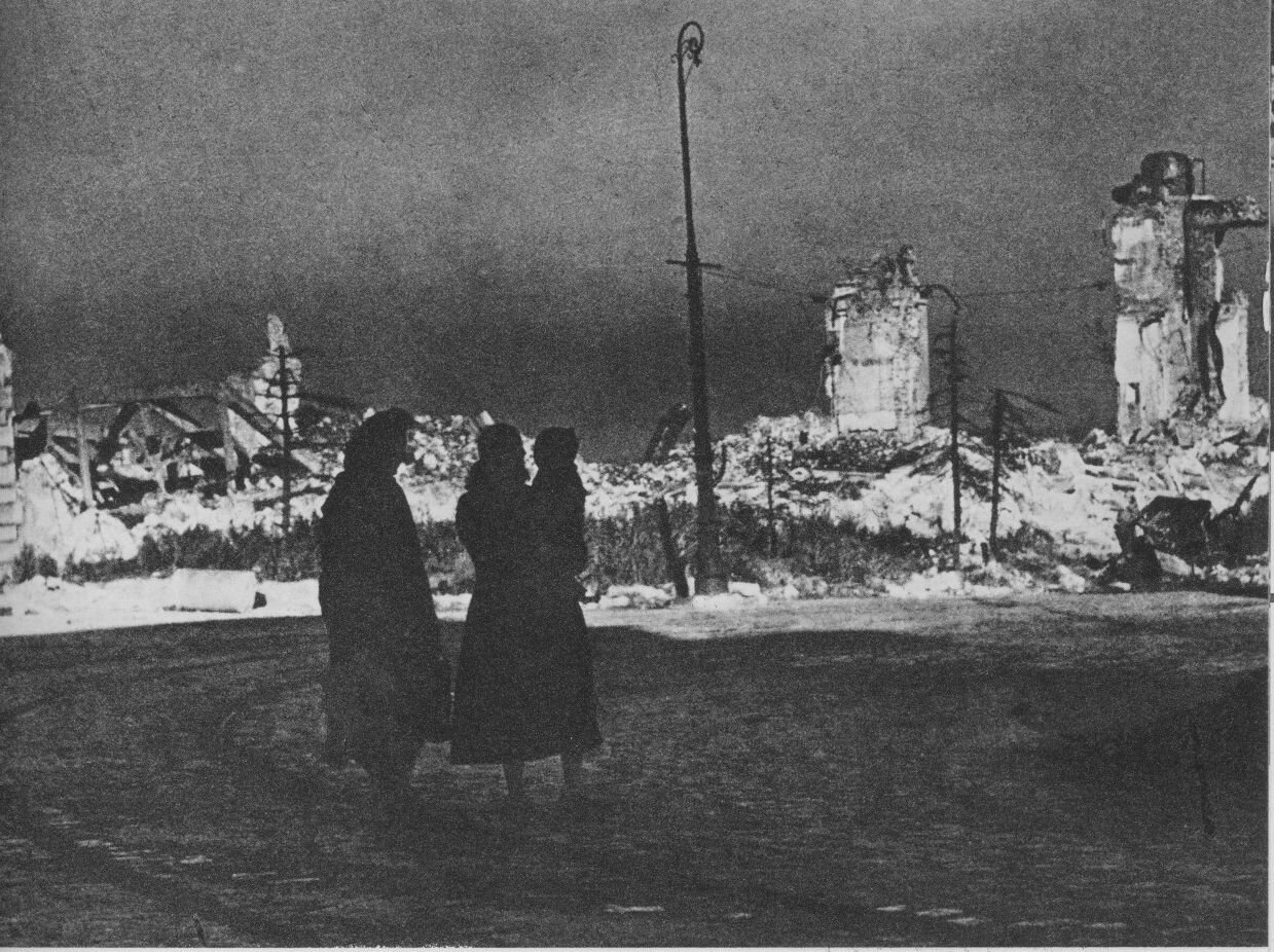 Zdjęcie przedstawia brukowaną ulicę, na której stoją dwie kobiety. Jedna z nich trzyma dziecko na rękach. Za ich plecami widoczne są gruzowiska. Nie ma ani jednego niezniszczonego budynku. Widać szczątki ścian i zwały cegieł oraz gruzu.