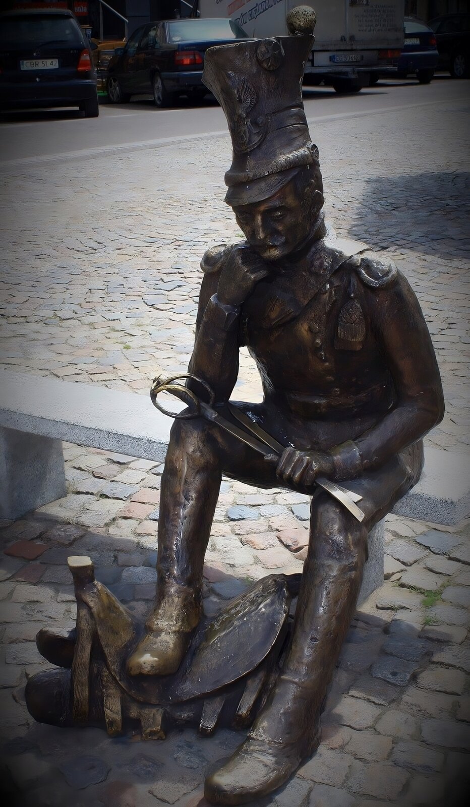 Zdjęcie przedstawia pomnik mężczyzny w mundurze armii napoleońskiej. Mężczyzna ubrany jest w obcisłe spodnie oraz marynarkę, do której przypięte zostały oznaczenia. Na głowie ma wysoką czapkę. Mężczyzna ma wąsy podkręcone na końcach. Prawą dłonią podpiera twarz, a w lewej trzyma złamaną na pół szablę. Prawą nogę postawił na siodle. Rzeźba jest brązowa. Postać siedzi na kamiennej ławce. W tle widoczne są zaparkowane samochody. Zdjęcie zostało wykonane w świetle dziennym. 