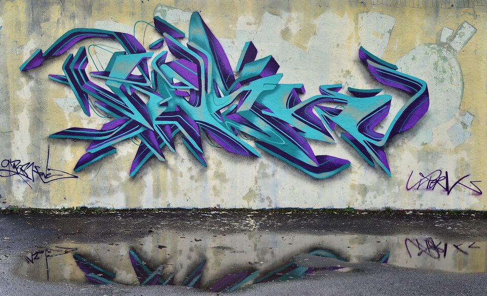 Zdjęcie przedstawia graffiti namalowane na jasnej ścianie. Graffiti ukazuje fioletowo - turkusowy napis. Pod graffiti jest kałuża, w której odbija się część grafiki.