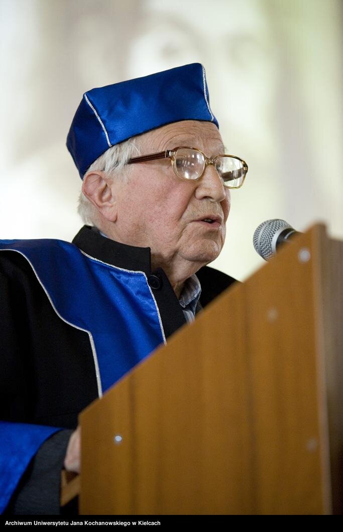 Zdjęcie przedstawia przemawiającego przy mównicy, starszego mężczyznę. Ma on siwe włosy, duże okulary na nosie. Ubrany jest w togę, na głowie ma biret.