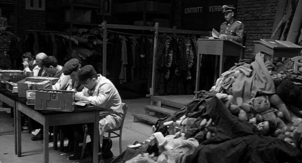 Zdjęcie przedstawia wnętrze fabrycznej hali. Na podwyższeniu, przy pulpicie stoi niemiecki oficer w mundurze. W hali leżą stosy ubrań. Przy stołach pracują mężczyźni.