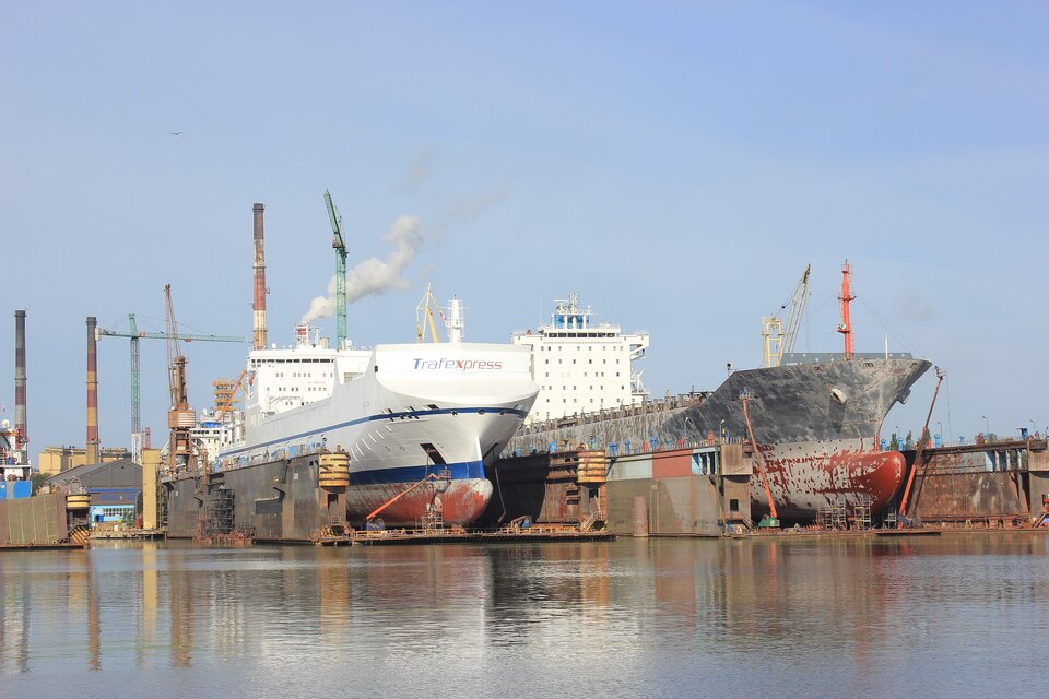 Zdjęcie przedstawia dwa duże statki stojące w stoczni. Za nimi są wysokie kominy, dźwigi i pagórek usypany z jakiegoś surowca. 
