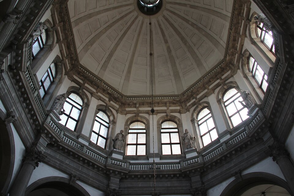 Widok w górę na wnętrze wysokiej, ośmiobocznej kopuły z promieniście podzielonym sklepieniem; poniżej biegnie galeria z balustradą, między dużymi półkolistymi oknami stoją kamienne rzeźby postaci, a światło wpada przez przeszklone okna, rozjaśniając jasne ściany i detale architektoniczne.