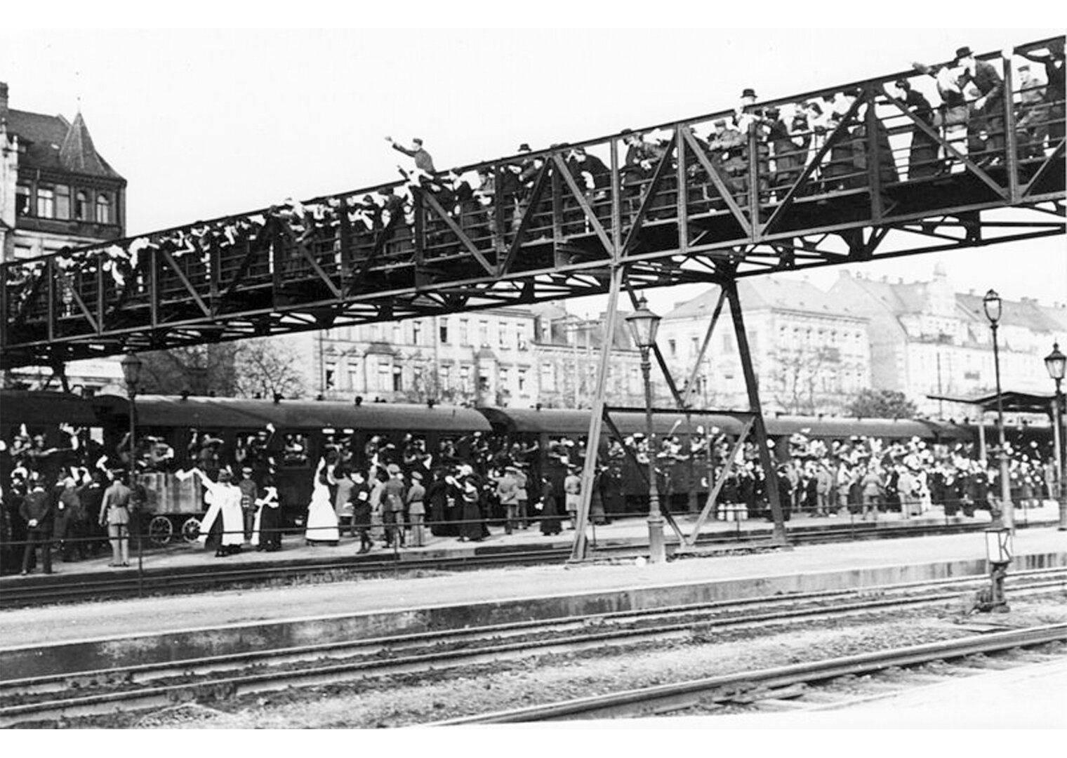 Fotografia czarno-biała przedstawiająca pociąg na stacji. Na moście nad torami kolejowymi oraz na peronie setki ludzi żegnających pasażerów pociągu. Machają im na pożegnanie. 