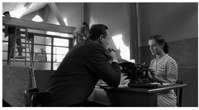 Zdjęcie przedstawia wnętrze fabryki. Robotnicy malują ściany. W sali stoi biurko, przy którym siedzi przy maszynie do pisania młoda kobieta o ciemnych włosach. Naprzeciw niej siedzi Schindler. Kobieta patrzy Schindlerowi w oczy. 