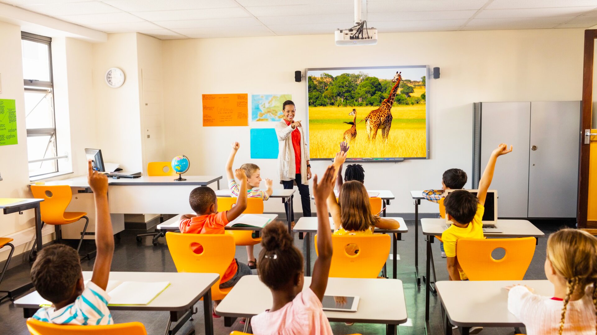 Ilustracja przedstawia salę lekcyjna z uczniami i nauczycielką z perspektywy widza, znajdującego się na końcu sali. Nauczycielka przedstawia materiały korzystając z tablicy multimedialnej. Na tablicy widać dwie żyrafy, prawdopodobnie matkę z dzieckiem. Uczniowie siedzą w pojedynczych ławkach. Zgłaszają się do odpowiedzi, podnosząc ręce. Być może jest to lekcja geografii.