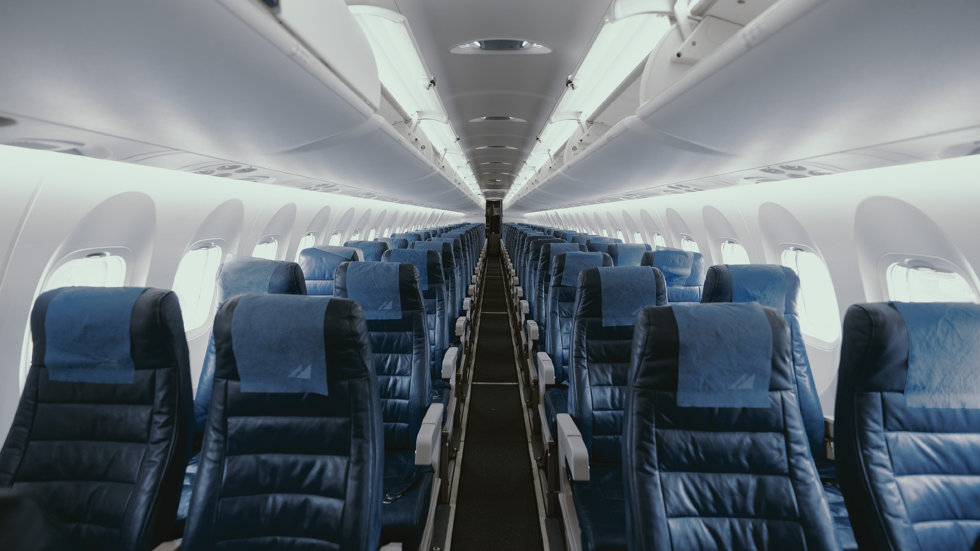 Zdjęcie przedstawia rzędy foteli na pokładzie samolotu pasażerskiego. W każdym rzędzie znajdują się cztery fotele, dwa po lewej i dwa po prawej stronie. Pośrodku znajduje się przejście. Widoczne są także okna w samolocie oraz otwarte półki na bagaże nad fotelami. Pokład jest pusty, nie ma pasażerów.