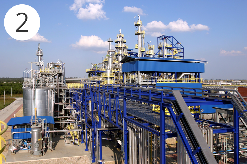 Zdjęcie przedstawia instalację przemysłową stosowaną w produkcji związków chemicznych w dużej skali. Jest ona złożona z szarych, jasnożółtych i niebieskich metalowych elementów. W tle znajduje się błękitne niebo z kilkoma białymi chmurami.