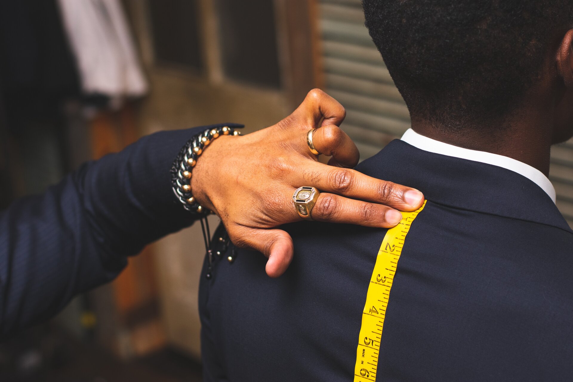 Zdjęcie przedstawia ciemnoskórego mężczyznę w garniturze, stojącego tyłem do widza. Kobieta mierzy długość jego pleców centymetrem.