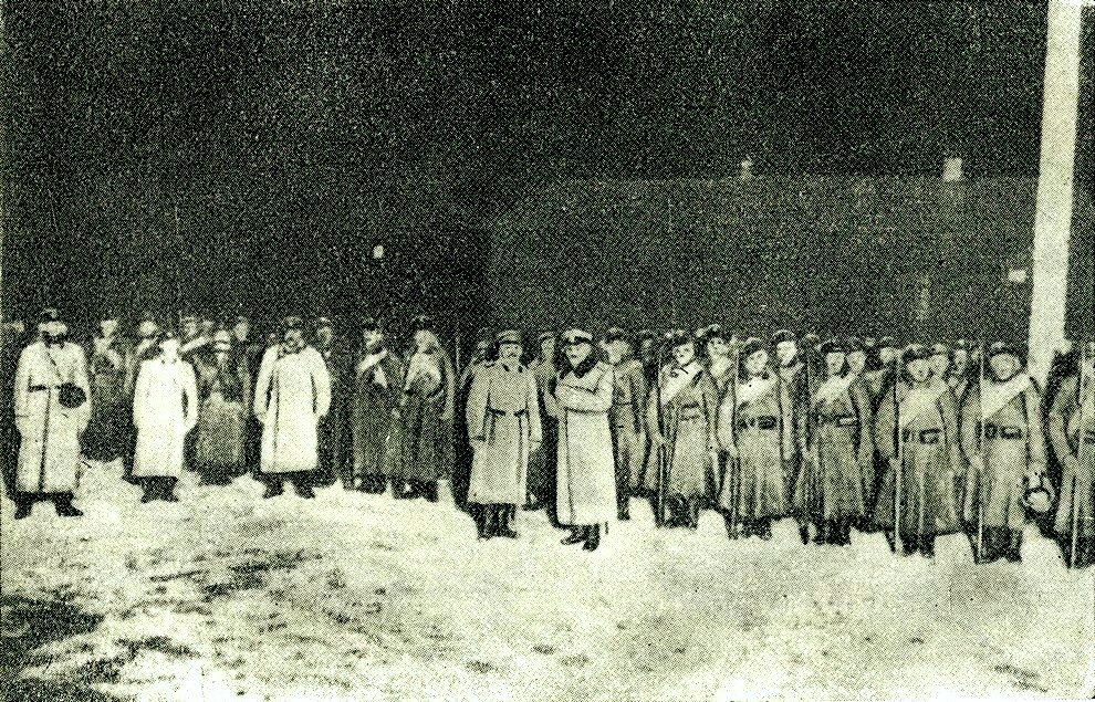 Zdjęcie przedstawia żołnierzy stojących w długich płaszczach i trzymających broń. 