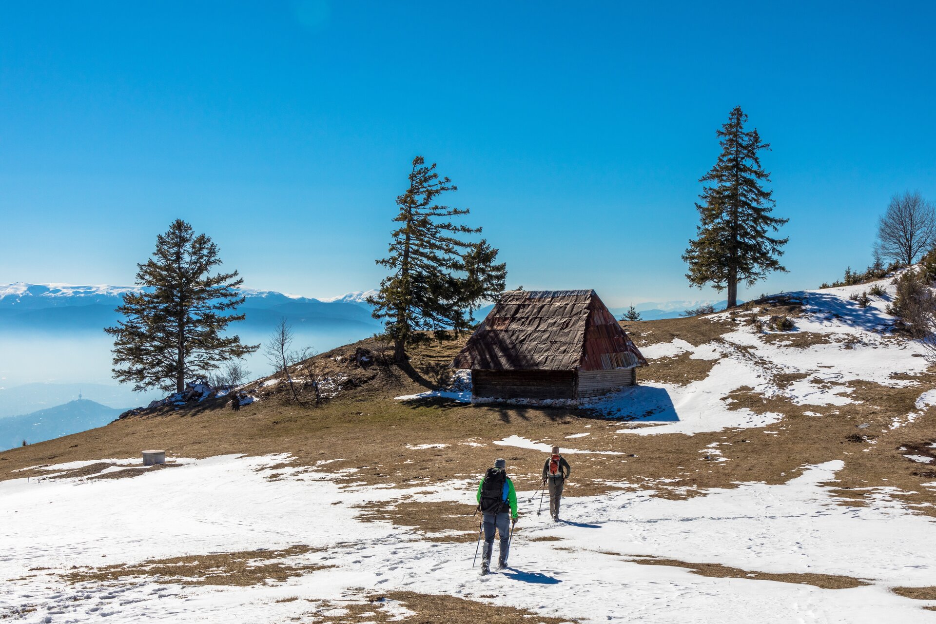 Widoczna jest fotografia. Przedstawia górską chatę i idące do niej dwie osoby. Chata znajduje się na szczycie, w tle widoczne są góry. Obok niej rosną drzewa, a w niektórych miejscach ziemię pokrywa śnieg. Widoczne na fotografii osoby prawdopodobnie spędzają wakacje w górach, wspinają się, wędrują, chodzą po lesie.