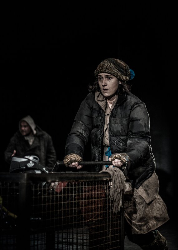Zdjęcie przedstawia kobietę w średnim wieku z ciemnymi włosami do ramion. Kobieta ma przerażony wzrok i lekko rozchylone usta. Ubrana jest w brązową kurtkę i brązową czapkę z niebieskim pomponem. Pcha przed sobą sklepowy wózek wypełniony rzeczami. W tle widać mężczyznę ubranego w płaszcz. Na głowę narzucił kaptur od bluzy.    