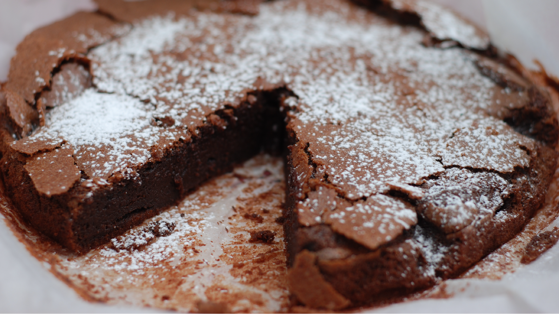 Na zdjęciu znajduje się okrągłe, włoskie ciasto czekoladowe o nazwie Tenerina, posypane cukrem pudrem. Jeden trójkątny kawałek z ciasta został już ukrojony.