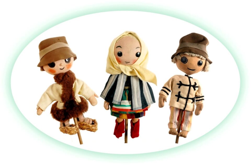 Zdjęcie przedstawia 3 teatralne lalki na patykach - tak zwane kukły. Lalki są niewielkich rozmiarów, mają okrągłe główki, oczy z przyszytych guzików, usta i brwi namalowane czarną, cienką kreską. Wszystkie są ubrane w polskie stroje ludowe. Pośrodku kukła dziewczynki ubrana jest w pasiastą spódnicę i krótką pelerynkę oraz białą chustkę na głowie, zawiązaną pod brodą. na nogach ma czerwone kozaki. Po dwóch stronach kukły chłopców w kapeluszach - z jednej strony, pastuszek w kubraczku obszytym futerkiem i słomianych łapciach na nogach, z drugiej strony, bosy chłopiec w odświętnym białym ubraniu.