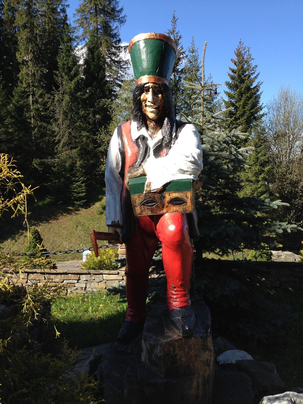 Kolorowa fotografia przedstawia rzeźbę harnasia Janosika, który nosi w czerwone spodnie i czerwoną kamizelkę oraz białą koszulę. Na głowie ma zielony kołpak czyli wysoką czapkę bez daszka w kształcie cylindra. Spod czapki wystają czarne włosy. Harnaś jedną nogę opiera na pniu drzewa, na kolanie trzyma kolorową zamkniętą szkatułę. W prawej dłoni ma siekierę. Rozbójnik uśmiecha się. W tle widać drzewa iglaste i zarys górskiego krajobrazu. 