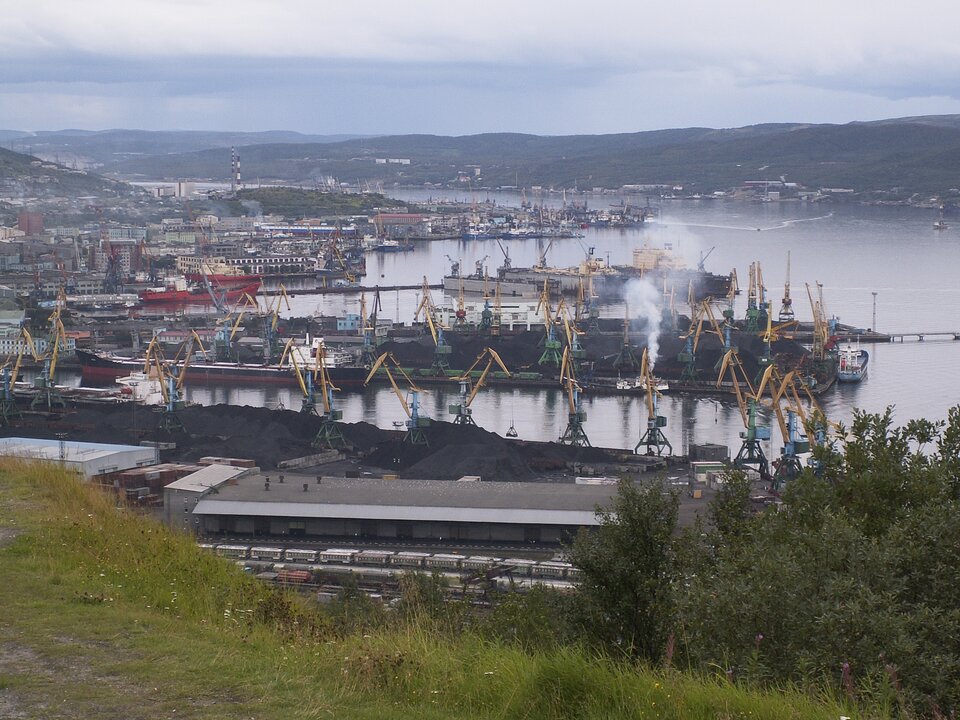 Zdjęcie przedstawia port w Murmańsku. W porcie stoją tankowce, są tam liczne dźwigi do załadunku surowca. Lewa strona brzegu portu jest gęsto zabudowana.  