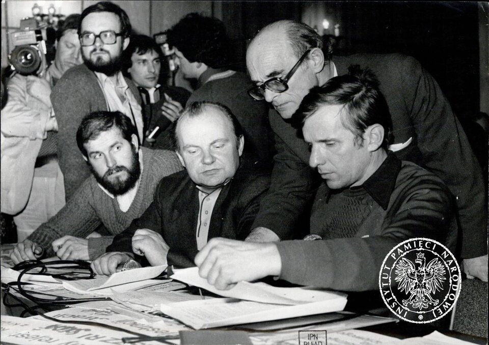 Ilustracja przedstawia działaczy chłopskiej „Solidarności" w Sądzie Najwyższym w Warszawie, w czasie drugiej rozprawy rewizyjnej o rejestrację. Mężczyźni są skupieni na dokumentach leżących na stole przed nimi. Czytają je, studiują. Za nimi znajdują się dziennikarze.