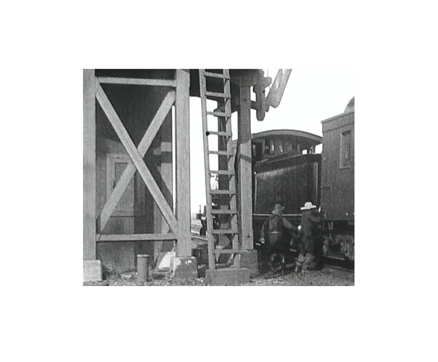 Kadr z czarno-białego filmu. Sytuacja dzieje się na dworze, najprawdopodobniej na dworcu kolejowym. Po prawej stronie kadru znajduje się pociąg, do którego wsiada dwoje mężczyzn w kowbojskich kapeluszach. Mężczyźni mogą być uzbrojeni. W centrum kadru na pierwszym planie znajduje się fragment budynku, drewniana konstrukcja z drabiną. Frame from black and white film. The situation is happening outside, most likely at the railway station. On the right side of the frame there is a train to which two men in cowboy hats get in. Men can be armed. In the center of the frame there is a fragment of the building in the foreground, a wooden structure with a ladder.