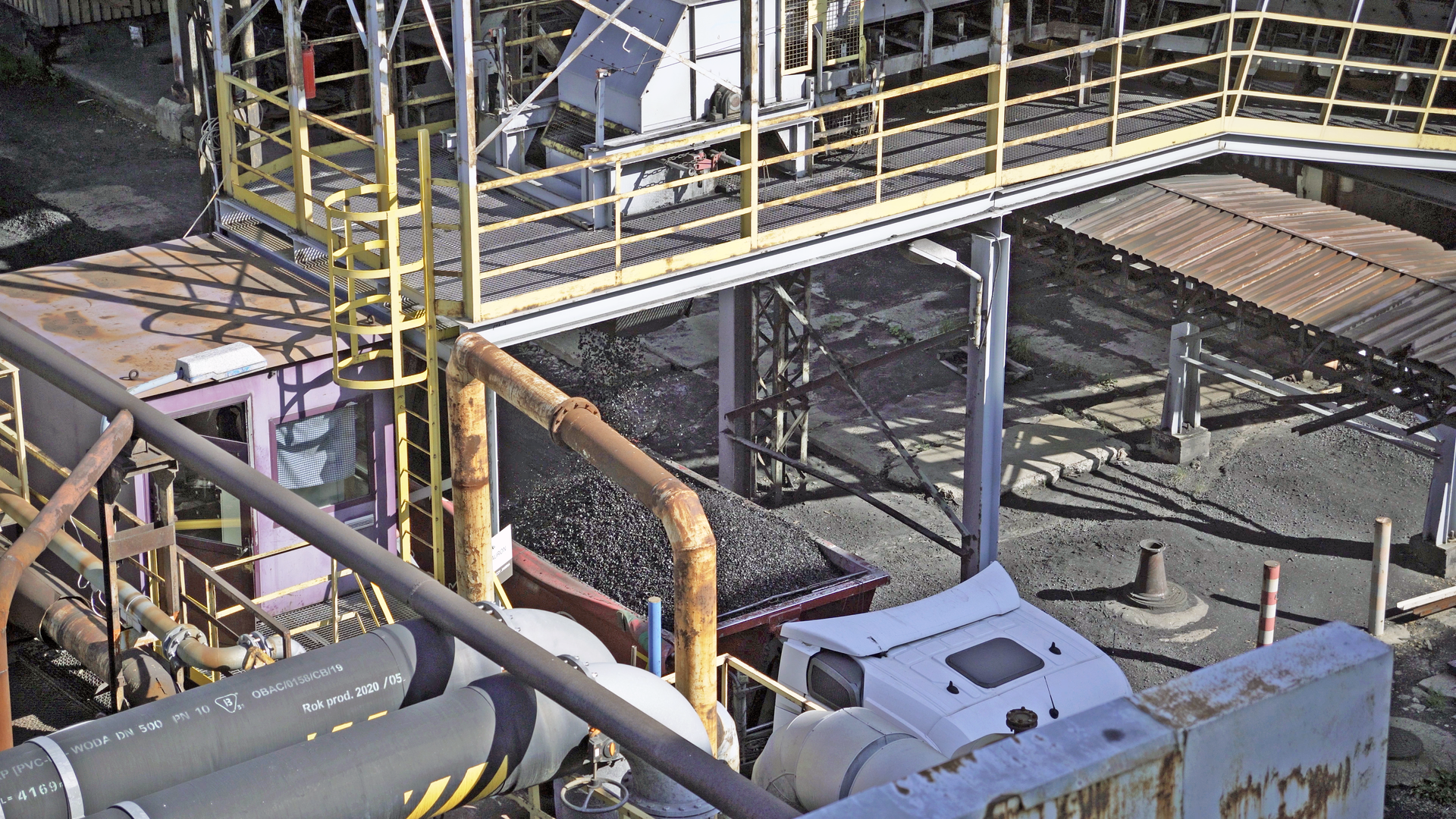 Na zdjęciu widoczny jest proces ładowania węgla na ciężarówkę. W centralnej części ekranu znajduje się ciężarówka. Naczepa ciężarówki jest w kształcie otwartego prostopadłościanu. W naczepie widoczny jest węgiel. Po lewej stronie ciężarówki znajduje się budynek z operatorem. Nad ciężarówką widać pomost. Pomost jest zabezpieczony żółta balustradą. .Poniżej zdjęcia widoczny jest przycisk multimedialny z nagraniem audio. Treść nagrania jest zgodna z tytułem zdjęcia.