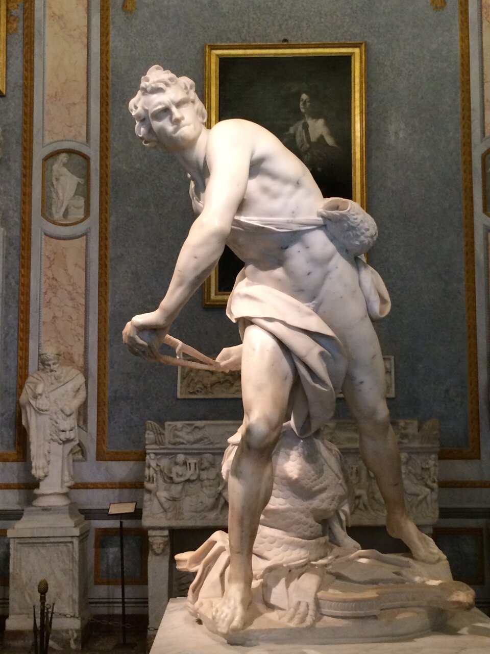 Ilustracja przedstawia rzeźbę „Dawid”, autorstwa Gianlorenzo Berniniego . Zdjęcie ukazuje marmurowy posąg nagiego, umięśnionego mężczyzny ustawionego w dynamicznej pozie. Postać biblijnego bohatera walczącego z Goliatem uchwycona została w momencie przygotowywania się do oddania rzutu. Prawie naga sylwetka z okrytymi jedynie skrawkiem materii biodrami oraz przewieszoną przez ramię torbą pasterską jest pełna napięcia. Dawid stoi w szerokim wykroku. Jego tułów skręcony jest w prawą stronę. Równolegle ustawione ręce trzymające naciągniętą procę z kamieniem odchylone są do tyłu za tułów. Głowa zwieńczona lokami włosów czujnie spogląda w przód. Twarz o zmarszczonym czole oraz zaciśniętych wargach wyraża skupienie. U stóp młodzieńca leży harfa oraz zbroja, którą zdjął przed walką. Artysta z mistrzostwem ukazał moment ruchu. Anatomia napiętego ciała oddana jest z niezwykłą precyzją.