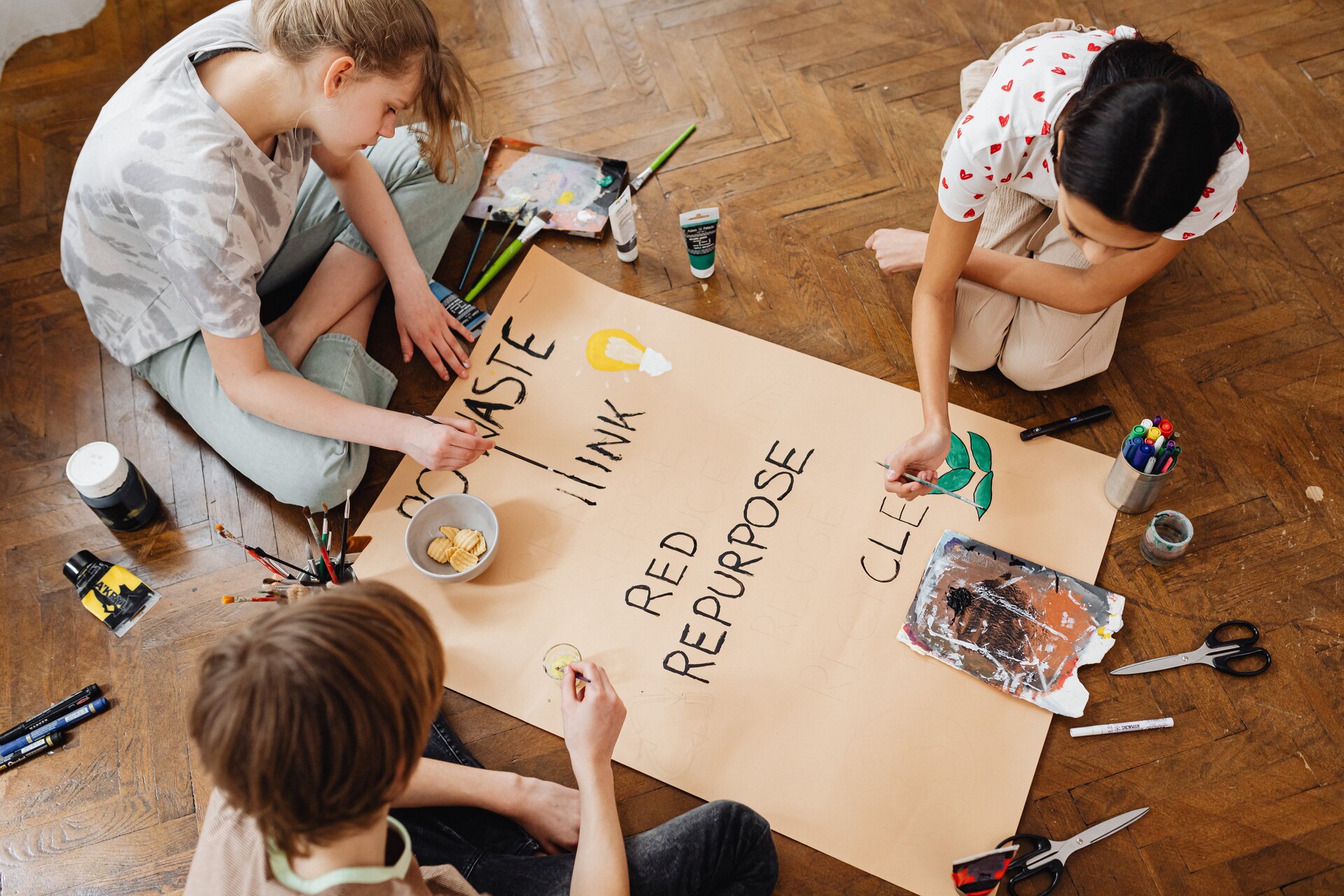 Ilustracja przedstawia trójkę dzieci, które siedzą na drewnianej podłodze i malują plakat. Dookoła nich leżą farby, nożyczki, taśmy klejące, kleje i inne niezbędne akcesoria do wykonania plakatu. 