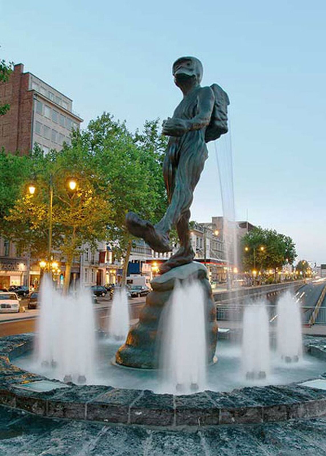 Ilustracja przedstawia fontannę w Brukseli wykonaną przez Luka van Soom pt. „Człowiek z Atlantydy”. Mężczyzna z plecakiem, który został uwieczniony w ruchu. Ma długie stopy i zniekształconą twarz.