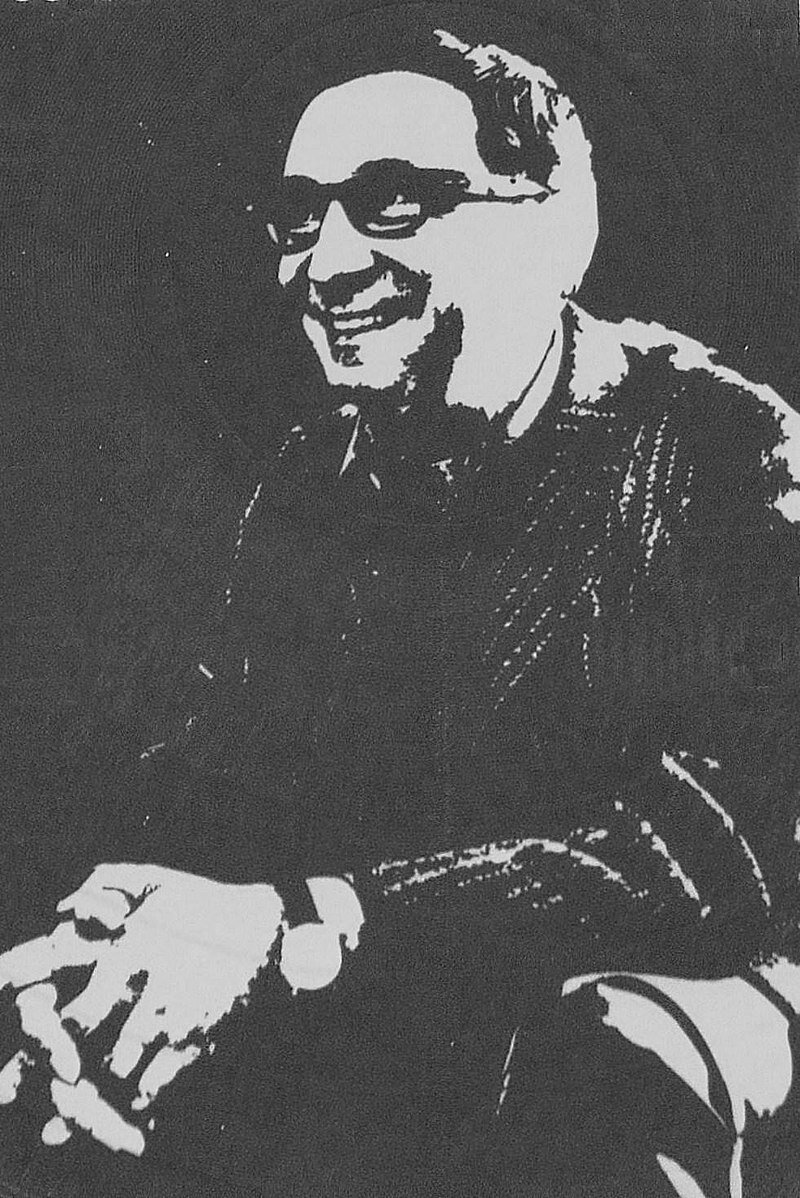 Rysunek przedstawia portret uśmiechniętego mężczyzny średnim wieku. Nosi okulary. Siedzi ze splecionymi dłońmi. Na lewej ręce ma założony zegarek.