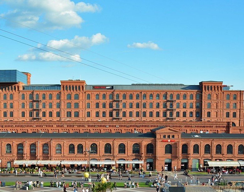 Długi, wielokondygnacyjny budynek z czerwonej cegły o regularnych rzędach okien i łukowych detalach architektonicznych stoi wzdłuż ruchliwego placu. Na parterze znajdują się lokale usługowe i restauracje z zadaszeniami, a przed budynkiem spacerują liczne osoby. Nad całością rozciąga się błękitne niebo z kilkoma chmurami, a przez kadr przebiegają linie napowietrzne.