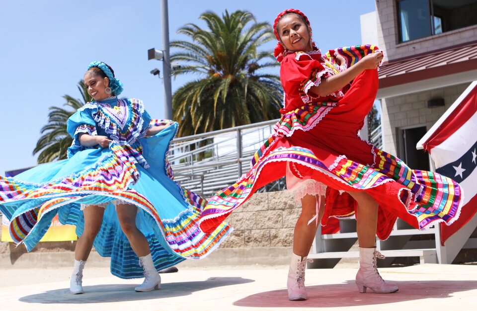 Zdjęcie przedstawia dwie tańczące kobiety. Mają na sobie, kolorowe długie suknie, ozdoby we włosach. W tle jest budynek oraz rosną palmy. 