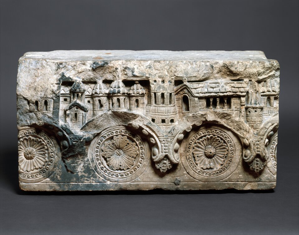 Romański kamienny relief architektoniczny: fragment fryzu z rytmicznymi rozetami i stylizowanymi arkadami, nad którymi przedstawiono miniaturowe budowle z wieżami i oknami; zachowany w surowej, jasno‑szarej kamiennej fakturze.