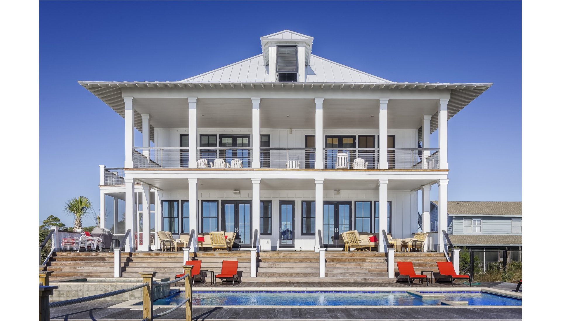 Zdjęcie przedstawia duży dwupoziomowy, luksusowy dom. Na pierwszym piętrze znajduje się duży taras. Taras zarówno na dole jak i na górze wsparty jest na kolumnach. Przed domem znajduje się basem. Przy basenie stoją leżaki.