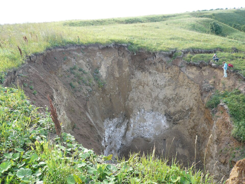  Ilustracja przedstawia lej krasowy. Jest to rozległe zagłębienie w ziemi w kształcie stożka głębokości nawet kilkudziesięciu metrów. Na jego brzegu widoczni są turyści, w oddali widać krzewy, trawy.