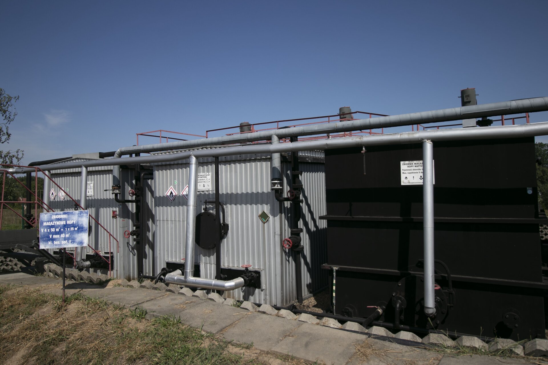Zdjęcie przedstawia zespół prostopadłościennych zbiorników magazynowych wraz z orurowaniem. Są one ze sobą połączone. Oznaczone piktogramami, wskazującymi na łatwopalność materiału w nich zawartego oraz jego szkodliwość dla zdrowia i środowiska. Na górze każdego z nich zamontowana jest platforma z barierką umożliwiająca poruszanie się pracownikom i dostęp do wnętrza zbiornika. Po lewej stronie znajduje się tabliczka. Na niebieskim tle biały napis. Zbiornik magazynowe ropy. <math aria‑label="fał">V cztery razy <math aria‑label="pięćdziesiąt metrów sześciennych">50 m3. Dodać jeden razy <math aria‑label="dwadzieścia pięć metrów sześciennych">25 m3. <math aria‑label="fał">V maksymalna <math aria‑label="czterdzieści metrów sześciennych">40 m3. Klasa bezpieczeństwa pożarowego pierwsza.