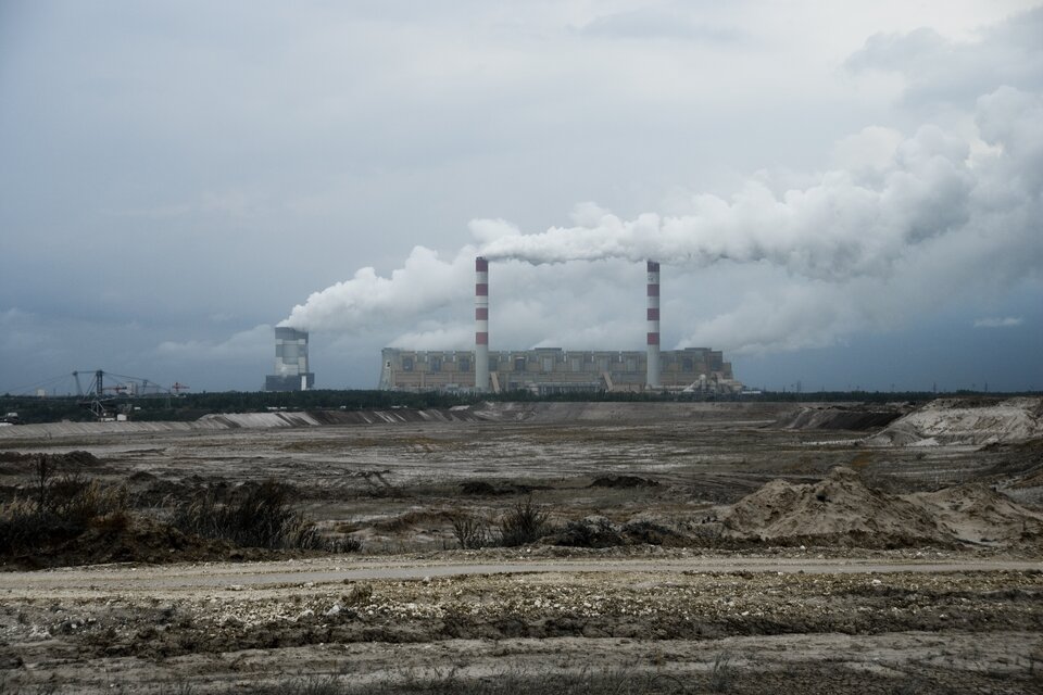 Zdjęcie przedstawia płaski teren pozbawiony roślinności. W tle jest elektrownia. Z kominów unosi się dym. 