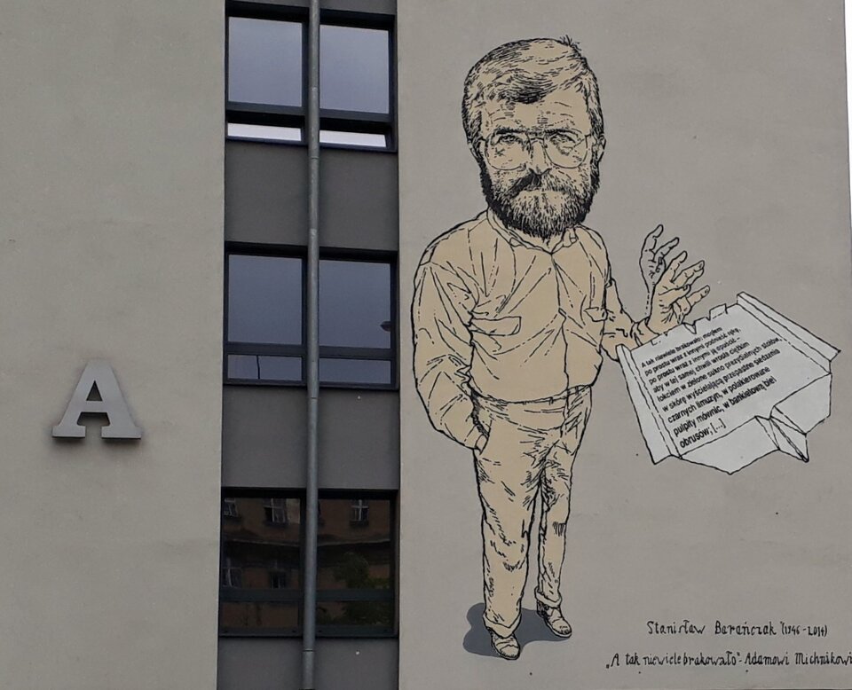 Zdjęcie przedstawia jasną ścianę, na której znajduje się mural przedstawiający mężczyznę. Ma on gęste włosy, wąsy oraz krótko przystrzyżoną brodę. Mężczyzna ma duże, okrągłe okulary. Ubrany jest w koszulę z długim rękawem oraz długie spodnie. Prawą dłoń włożył do kieszeni spodni, a lewą zgiął w łokciu i unosi ku górze. Przy jego ręce widoczny jest list, na którym są nieczytelne zapiski. Pod muralem znajduje się podpis - "Stanisław Barańczak (1946 - 2014) 'A tak niewiele brakowało' Adamowi Michnikowi". Mural umieszczony został obok czarno‑białej szyby ciągnącej się przez całą wysokość budynku. Po drugiej stronie okien widoczna jest wypukła litera "A". 