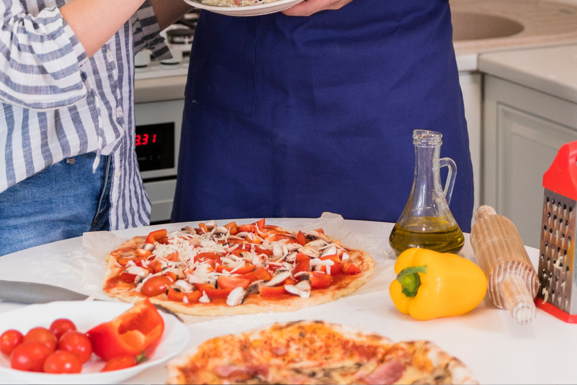 Zdjęcie przedstawia dwie osoby, widziane tylko na wysokości pasa, stojące przy okrągłym blacie kuchennym. Osoba po prawej ma przewiązany w pasie fartuch kuchenny. Na blacie widoczne są: biały głęboki talerz z małymi pomidorami i przekrojoną czerwoną papryką, przygotowana do wypieku pizza, fragment upieczonej pizzy, żółta papryka, mała szklana karafka z oliwą, drewniany wałek i tarka.