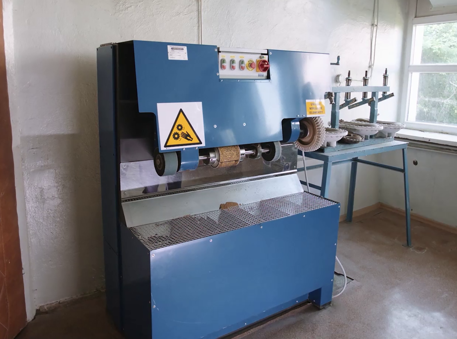 Zdjęcie pod tytułem: Czyszczarka. Zdjęcie przedstawia dużą, stojącą na podłodze maszynę z włącznikiem w górnej części, blatem z metalowej siatki i czterema szczotkami w formie tarcz z różnych materiałów. 