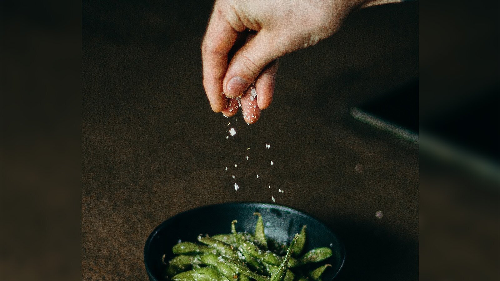 Zdjęcie przedstawia przyprawianie potrawy solą. U góry widoczna jest prawa dłoń, z której sypią się kryształki białej soli i spadają na zielony groszek znajdujący się w czarnej miseczce.