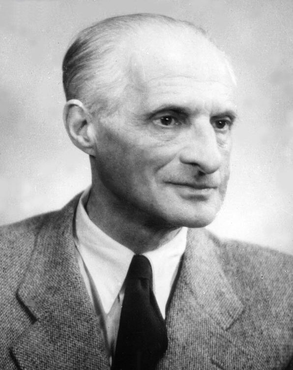 Zdjęcie przedstawia portret starszego mężczyzny. Ukazany jest prawy półprofil mężczyzny. Jego włosy są rzadkie, krótkie, zaczesane do góry. Mężczyzna ma pociągłą twarz ze zmarszczkami. Ma głęboko osadzone oczy. Jego nos jest wydatny. Ubrany jest w garnitur, koszulę i krawat.