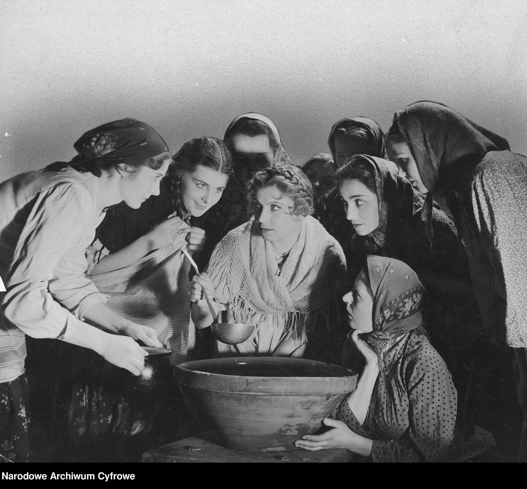 Fotografia przedstawiająca grupę młodych kobiet pochylających się nad ceramiczną miską. Większość z nich ma na głowach zawiązane chustki. Ubrane są w koszule. Kobieta stojąca pośrodku ma gołą głowę ozdobioną warkoczem upiętym wokół niej. Patrzy w lewą stronę. Ukazuje lewy profil. Zaprezentowana jest od pasa w górę. Kobieta ma owalną twarz, cienkie brwi, duże, okrągłe oczy, prosty nos i wydatne usta. Kobieta ma zawieszony przez ramiona szal wykonany na drutach. W prawej dłoni trzyma metalową chochlę.